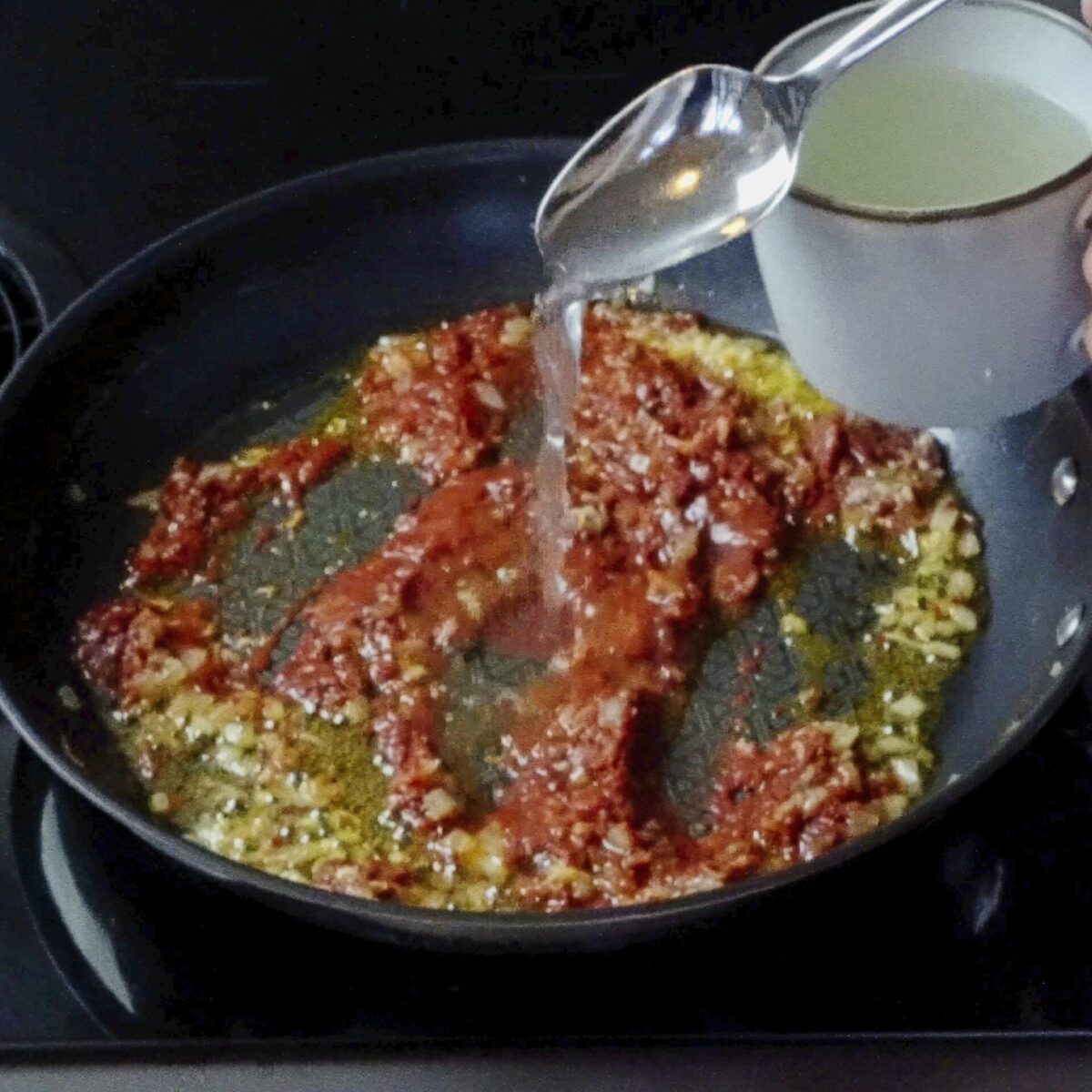 Pouring water into tomato paste in the pan to create the sauce base.