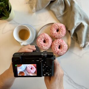 A person holding a camera, taking a photo of donuts with pink glaze.