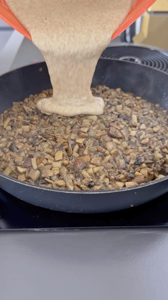 Pouring batter onto sautéed mushrooms in a pan.