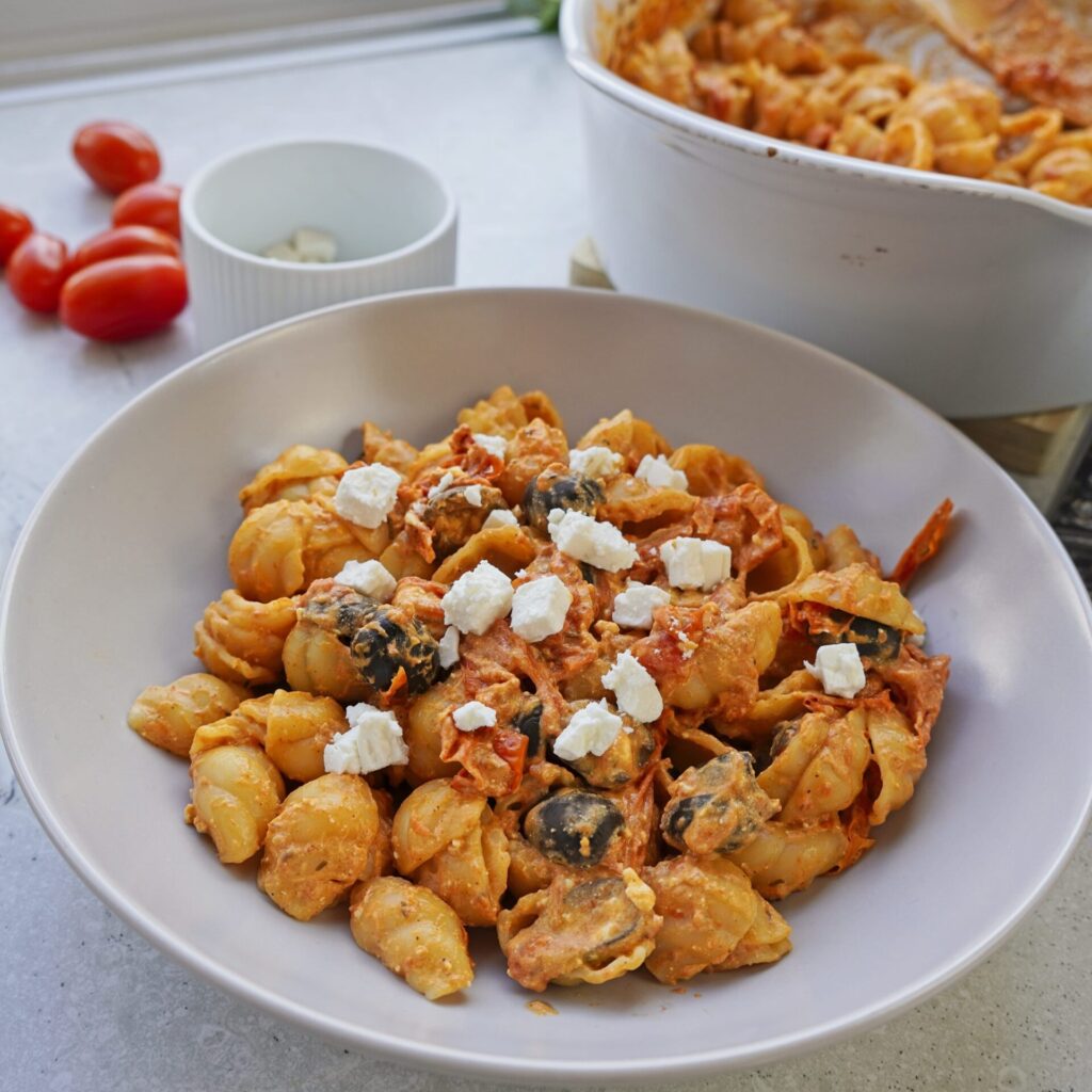 A plate of pasta with tomato and feta cheese on a table, with a baking form of freshly baked pasta in the background.