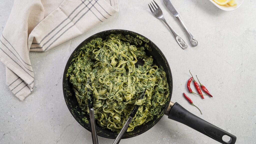 A pan full of pasta with ricotta and spinach sauce. The pasta looks creamy and smooth. A fork and knife are near a pan, ready for a person to start eating. 