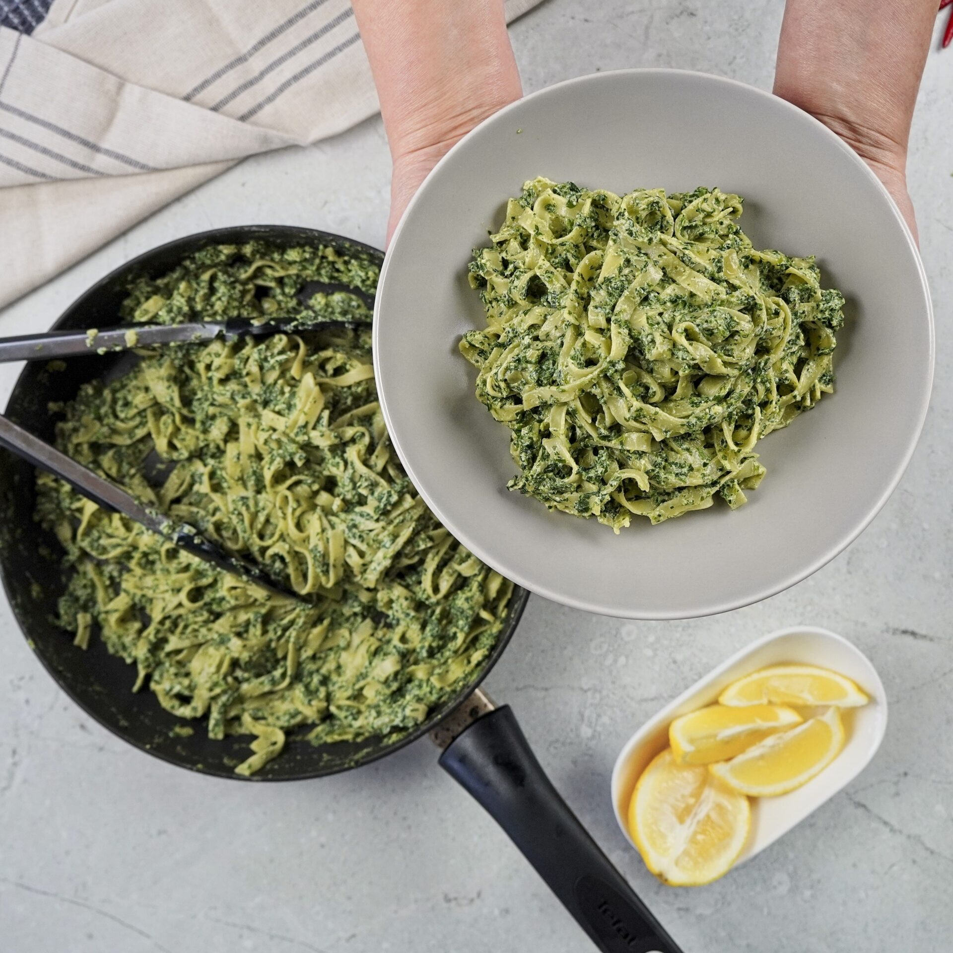 A person holding a plate with ricotta and cheese pasta. Pasta sauce looks creamy, smooth, and very delicious. In the background, there is a pan with more pasta.