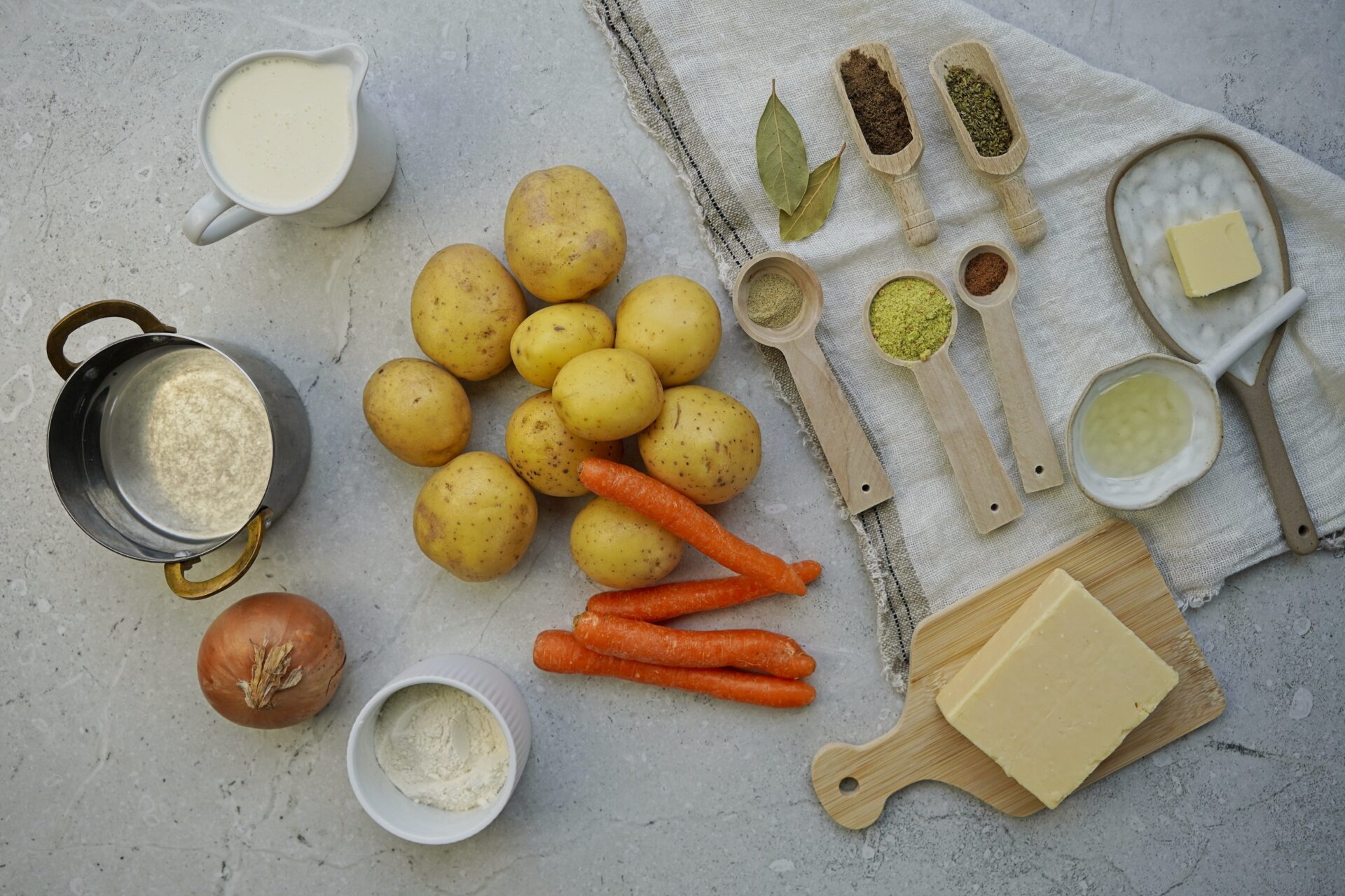 Ingredients you need for potato cheese soup: potatoes, neutral vegetable oil, butter, onion, carrot, dry vegetable bouillon, ground cumin, oregano, white pepper, nutmeg, bay leaves, heavy cream, flour, and cheddar cheese.