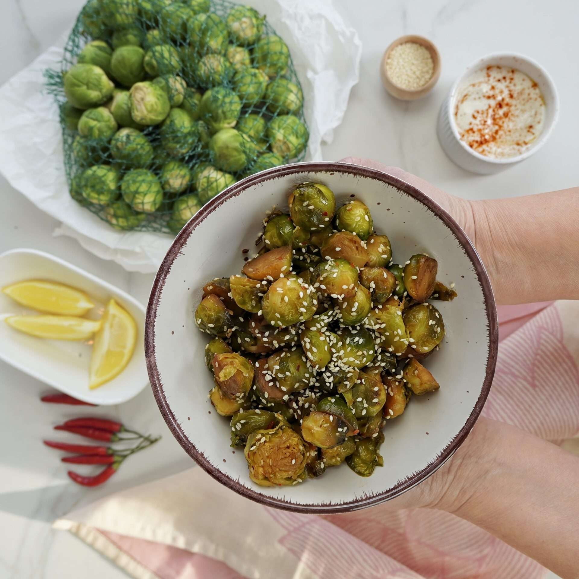 Brussels sprouts on a plate. On top, there are sesame seeds. On the right side is yogurt dip, and on the left, raw Brussels sprouts are in a net bag.